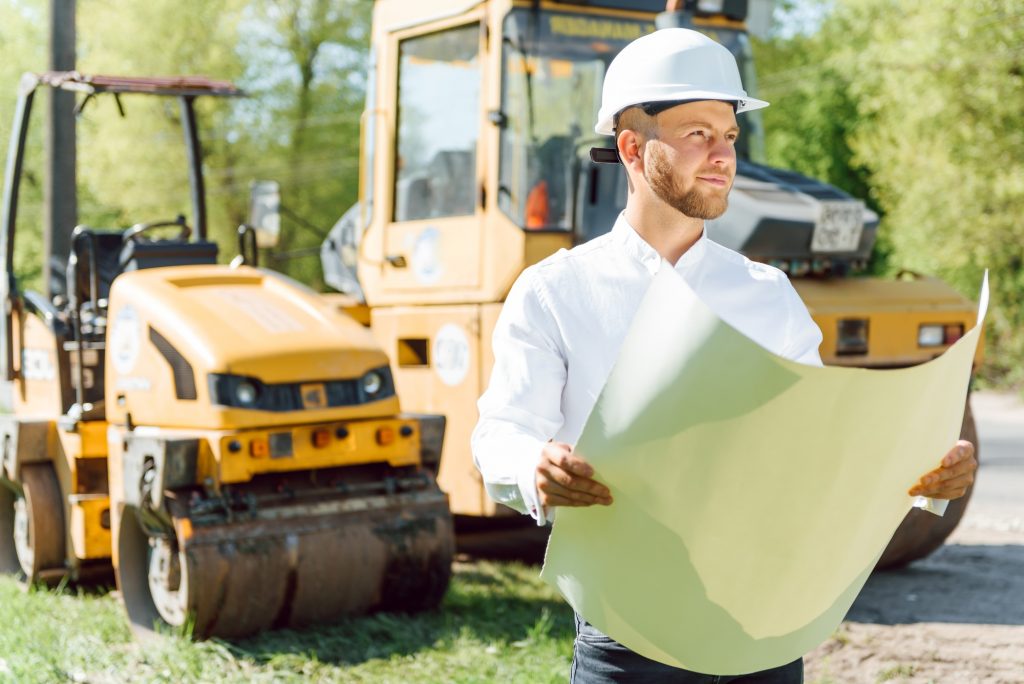 Smiling engineer with helmet standing in front of excavator on road construction site